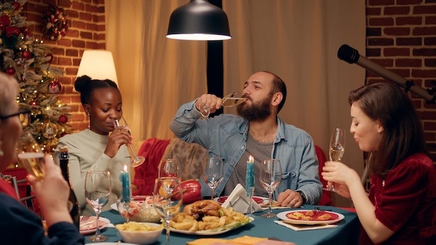 Pessoas diversas festivas desfrutando de reencontro enquanto celebram as férias de inverno com comida tradicional e álcool. Membros da família felizes brindando com champanhe enquanto estão sentados à mesa de jantar de Natal em casa