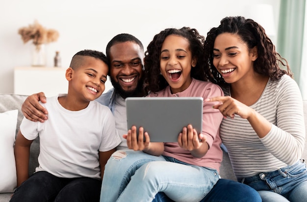 Pessoas e Tecnologia. Retrato de família afro-americana feliz segurando e usando computador tablet digital sentado no sofá em casa. Pessoas assistindo filmes, passando tempo juntos e rindo