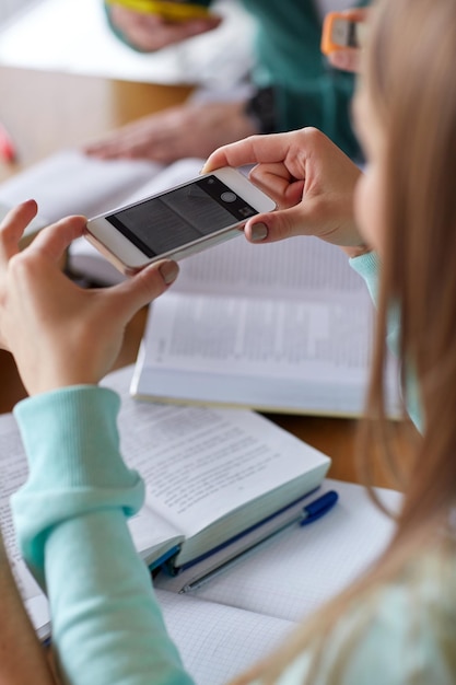 Pessoas, educação, tecnologia e conceito de exame - close-up das mãos do aluno com smartphone tirando foto da página de livros e fazendo cola na biblioteca escolar