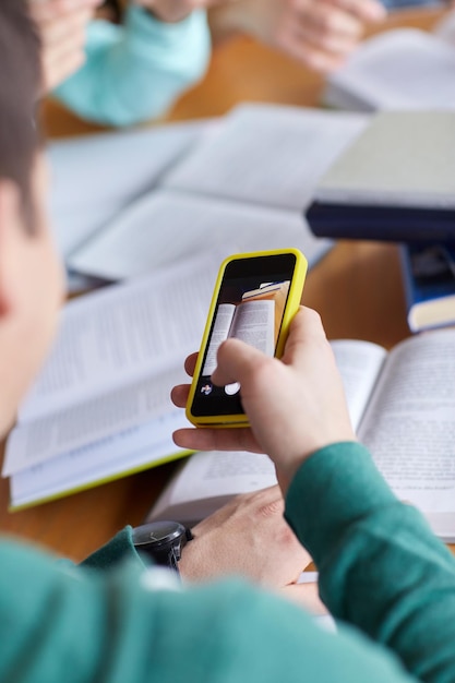 pessoas, educação, tecnologia e conceito de exame - close-up das mãos do aluno com smartphone tirando foto da página de livros e fazendo cola na biblioteca escolar