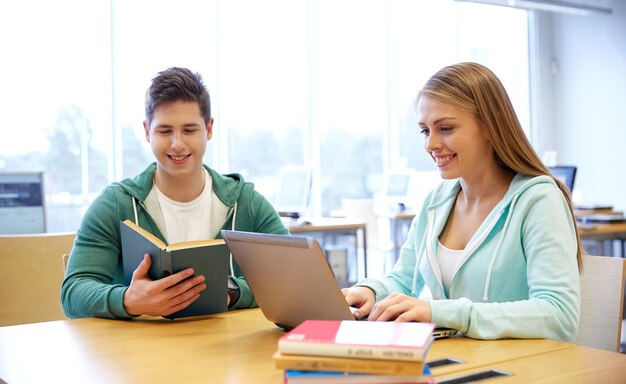 pessoas, educação, tecnologia e conceito escolar - estudantes felizes com computador portátil e livros na biblioteca