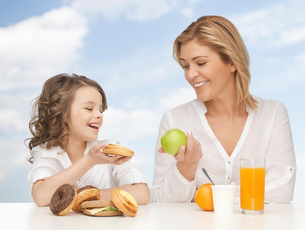 pessoas, estilo de vida saudável, conceito de família e comida - feliz mãe e filha tomando café da manhã saudável sobre fundo de céu azul