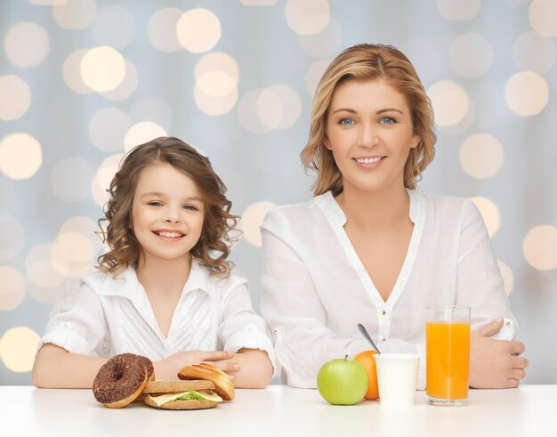 Pessoas, estilo de vida saudável, família e conceito de comida não saudável - feliz mãe e filha comendo comida diferente sobre fundo de luzes de férias