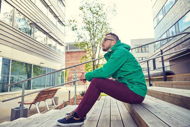 pessoas, estilo, lazer e estilo de vida - feliz jovem hipster em óculos e jaqueta sentado nas escadas na cidade