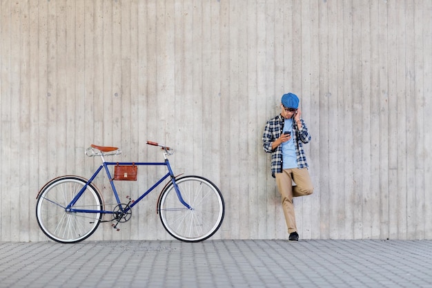 pessoas, estilo, tecnologia, lazer e estilo de vida - homem jovem hipster em fones de ouvido com smartphone e bicicleta fixa ouvindo música na parede da rua da cidade