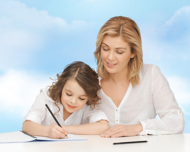 pessoas, família, escola, educação em casa e conceito de paternidade - feliz mãe e filha fazendo lição de casa e escrevendo no caderno sobre fundo de céu azul