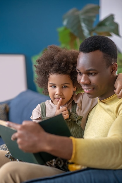 Pessoas felizes. afro-americano lendo um livro interessante para sua filhinha atenta e abraçada, sentada no sofá em casa