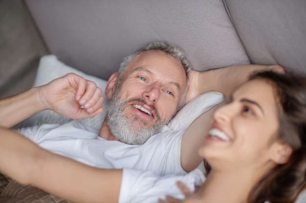 Pessoas felizes. Close de rostos felizes de um homem barbudo de cabelos grisalhos e uma mulher entusiasmada de cabelos escuros, deitados na cama