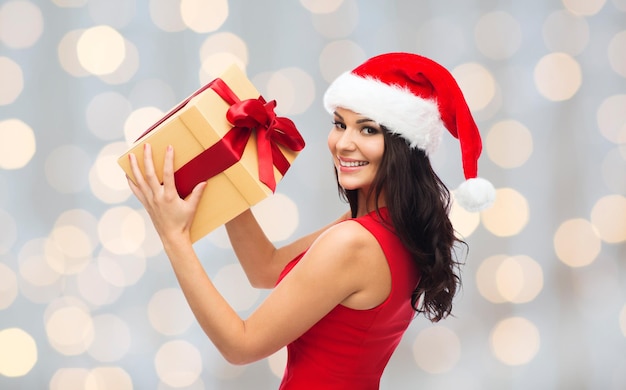 pessoas, feriados, natal e conceito de celebração - linda mulher sexy de vestido vermelho e chapéu de papai noel com caixa de presente sobre fundo de luzes de feriados