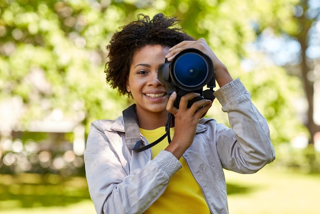 pessoas, fotografia, tecnologia, lazer e estilo de vida - feliz jovem fotógrafa afro-americana olhando para o visor da câmera digital no parque de verão