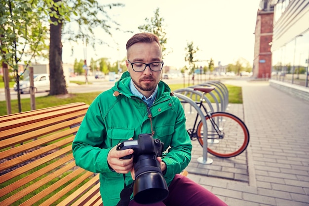 pessoas, fotografia, tecnologia, lazer e estilo de vida - jovem hipster segurando e olhando para câmera digital com lente grande na rua da cidade