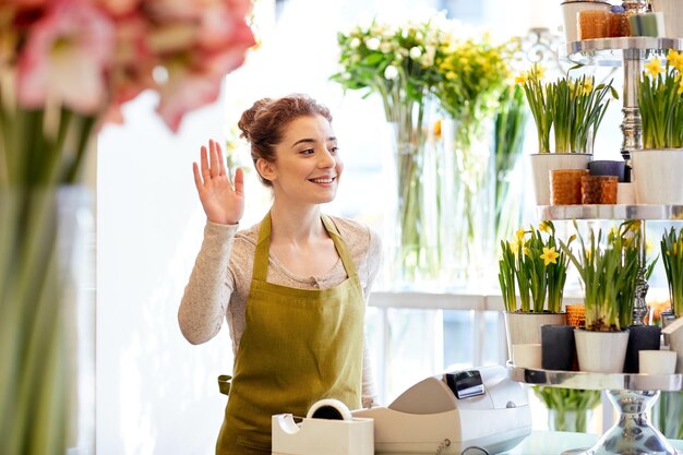 Foto pessoas, gesto, negócios, venda e conceito de floricultura - mulher florista sorridente feliz acenando com a mão no caixa da loja de flores