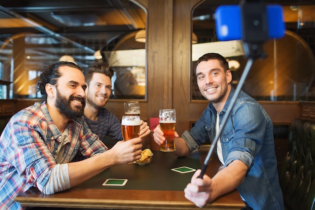 Foto pessoas, homens, lazer, amizade e conceito de tecnologia - amigos do sexo masculino felizes bebendo cerveja e tirando foto com smartphone selfie stick no bar ou pub