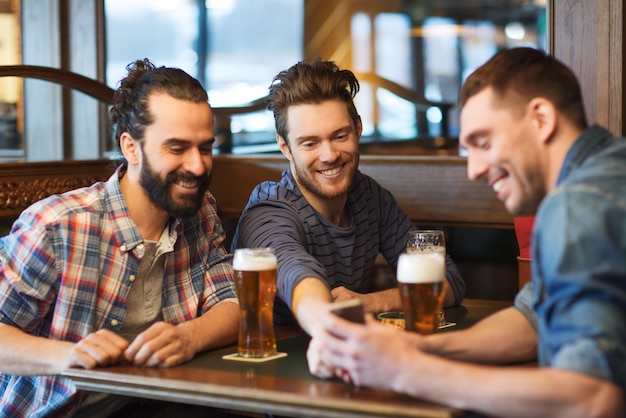 pessoas, homens, lazer, amizade e conceito de tecnologia - amigos do sexo masculino felizes com smartphone bebendo cerveja no bar ou pub