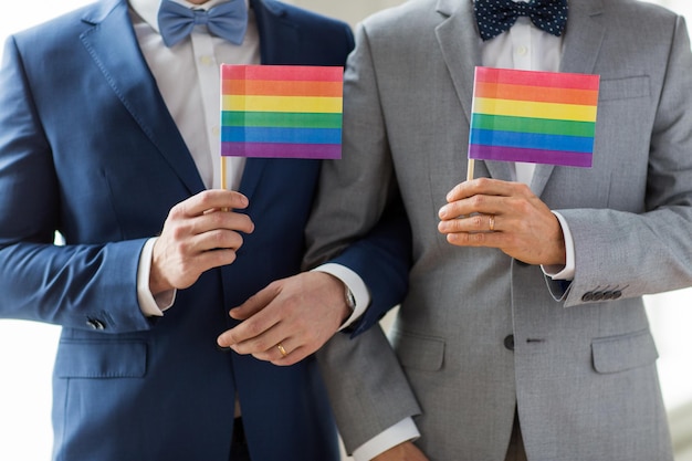 Foto pessoas, homossexualidade, casamento do mesmo sexo e conceito de amor - close-up de feliz casal gay masculino de terno e gravata borboleta com anéis de casamento segurando bandeiras do arco-íris