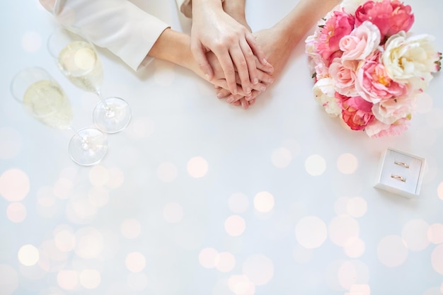 pessoas, homossexualidade, casamento do mesmo sexo e conceito de amor - close-up de mãos de casal de lésbicas feliz com bando de flores, taças de champanhe e anéis de casamento sobre fundo de luzes de férias