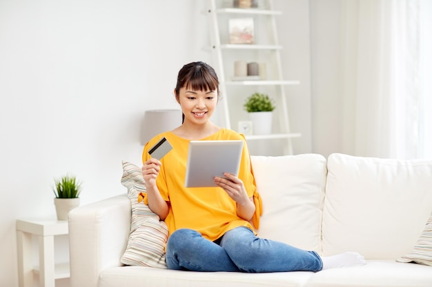 pessoas, internet bank, compras online, tecnologia e conceito de dinheiro eletrônico - jovem asiática feliz sentada no sofá com computador tablet pc e cartão de crédito em casa