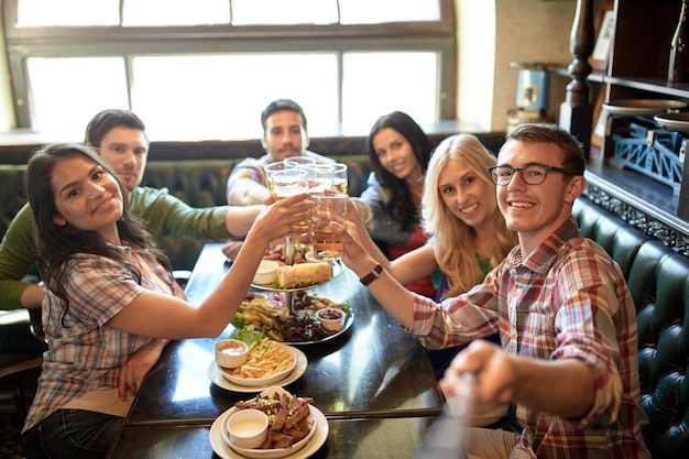 pessoas, lazer, amizade e conceito de tecnologia - amigos felizes tirando fotos com bastão de selfie, bebendo cerveja e comendo lanches no bar ou pub