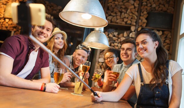 pessoas, lazer, amizade, tecnologia e conceito de comunicação - grupo de amigos sorridentes felizes com smartphone no pau de selfie e bebidas tirando foto no bar ou pub