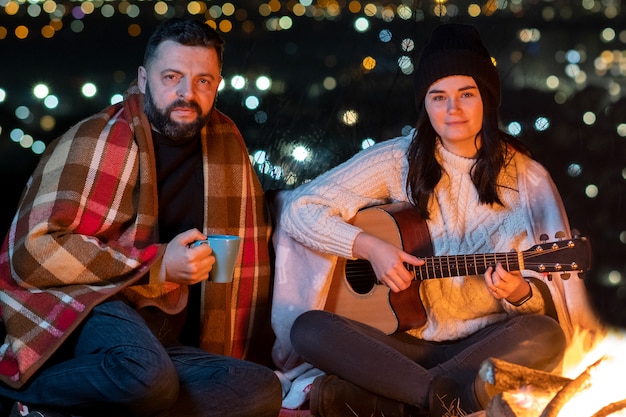 Pessoas se divertindo sentado perto da fogueira ao ar livre à noite tocando violão