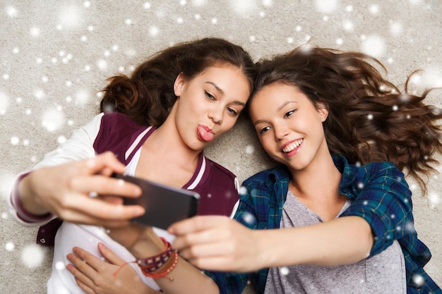 Foto pessoas, tecnologia, inverno, natal e conceito de amizade - felizes sorrindo muito adolescentes ou amigos deitados no chão e tirando selfie com smartphone sobre a neve