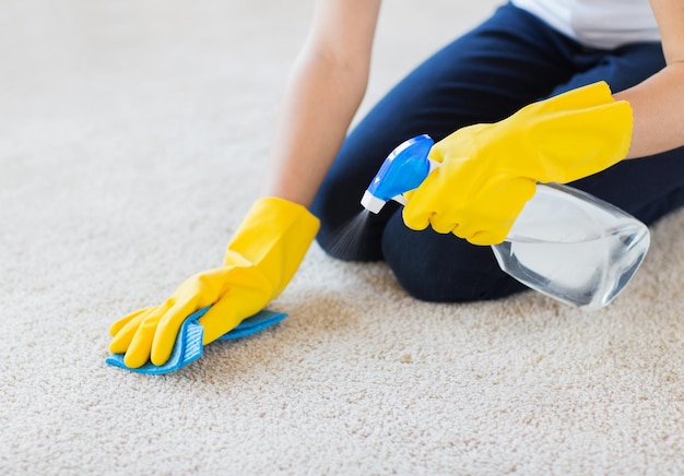 pessoas, trabalho doméstico e conceito de limpeza - close-up de mulher em luvas de borracha com pano e detergente spray limpeza tapete em casa