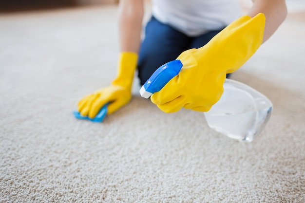 pessoas, trabalho doméstico e conceito de limpeza - close-up de mulher em luvas de borracha com pano e detergente spray limpeza tapete em casa