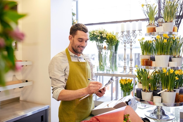 pessoas, venda, varejo, negócios e conceito de floricultura - florista sorridente feliz com cacho escrevendo e fazendo anotações na área de transferência no balcão da floricultura