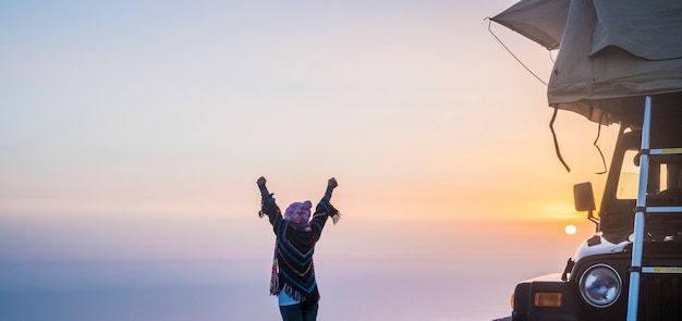 Pessoas viajam sozinhas com carro e barraca para um estilo de vida de acampamento grátis - alternativa de férias férias com uma mulher apreciando o pôr do sol do lado de fora do veículo em frente a um céu colorido com sol