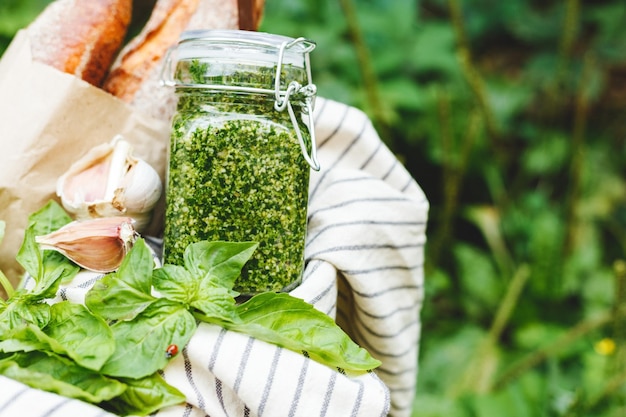 Pesto caseiro em uma jarra de vidro com manjericão, alho, pão ao ar livre em uma cesta. cesta de piquenique com molho pesto para preparar pão de alho para bruschetta.molho italiano tradicional - pesto. copie o espaço.