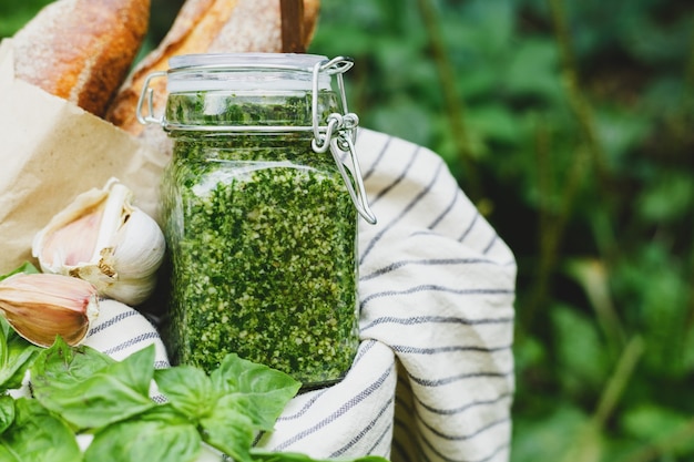 Pesto casero en un frasco de vidrio con albahaca, ajo, pan al aire libre en una canasta. Cesta de picnic con salsa Pesto para preparar pan de ajo para bruschetta. Salsa tradicional italiana - Pesto. Copie el espacio.