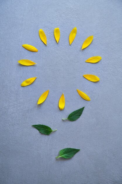 Pétalas amarelas colocadas em uma forma circular simulando uma flor com folhas verdes no centro da imagem de cima em um fundo listrado de textura cinza com luz natural