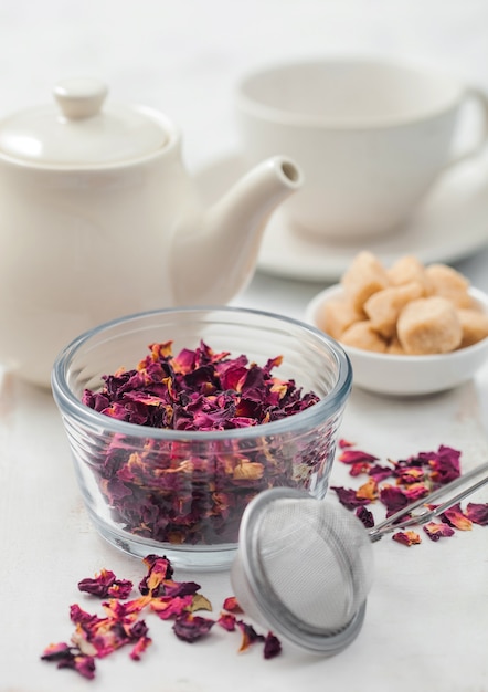 Pétalos de rosa orgánicos frescos con infusor de colador y tetera de cerámica blanca y azúcar de caña sobre fondo blanco.