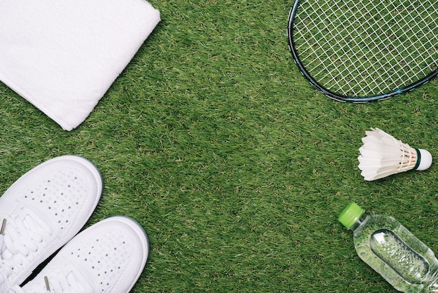 Peteca e raquete de badminton com sapato, água e toalha no fundo da grama.