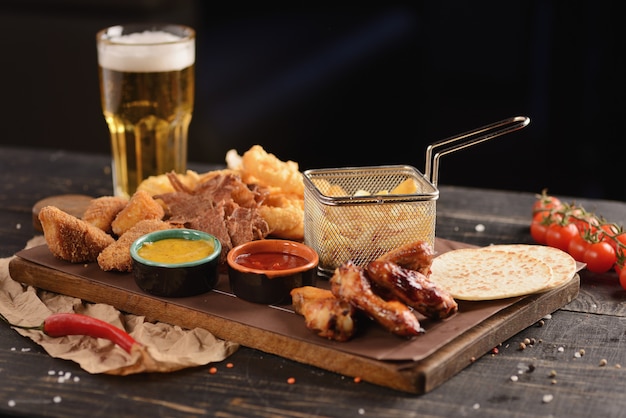 Foto petiscos de cerveja. asas de frango frito, batata frita, rodelas de cebola, queijo em massa e carne seca. em uma placa de madeira