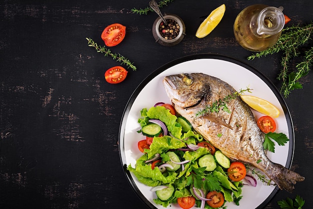 Pez dorado al horno con limón y ensalada fresca en plato blanco sobre un fondo rústico oscuro Vista superior Cena saludable con concepto de pescado Dieta y alimentación limpia