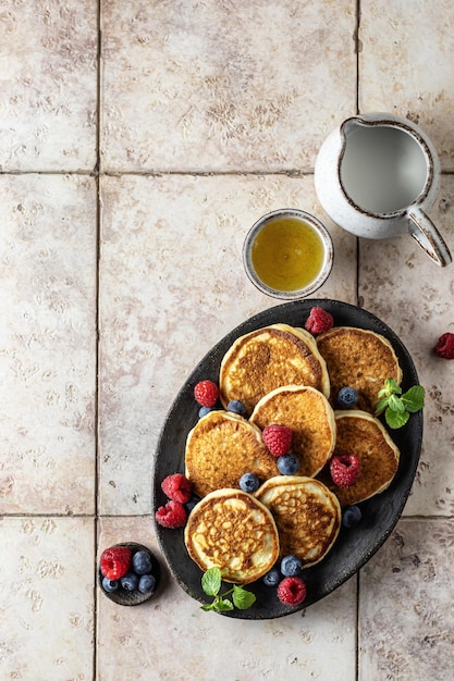 Pfannkuchen, Himbeere, Heidelbeere und Honig auf ovaler Platte auf rosafarbenem Fliesenhintergrund mit Textraum, flache Frühstückskomposition