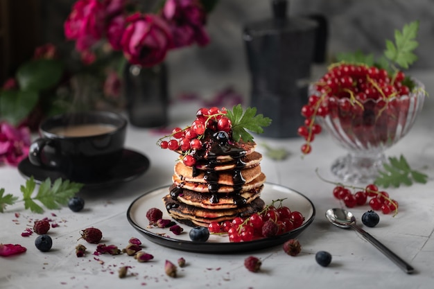 Pfannkuchen mit Beeren und Schokolade auf hellem Hintergrund mit einem Strauß roter Rosen. Dunkles Foto.