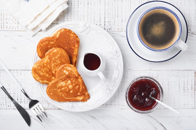 Pfannkuchen mit Beerenmarmelade und Honig in Form von Herzen und heißer Tasse Kaffee auf weißem Holzhintergrund. Konzeptfrühstück zum Valentinstag oder angenehme Überraschung für einen geliebten Menschen Tisch von oben betrachtet