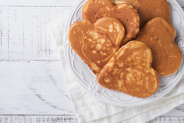 Pfannkuchen mit Beerenmarmelade und Honig in Form von Herzen und heißer Tasse Kaffee auf weißem Holzhintergrund. Konzeptfrühstück zum Valentinstag oder angenehme Überraschung für einen geliebten Menschen Tisch von oben betrachtet