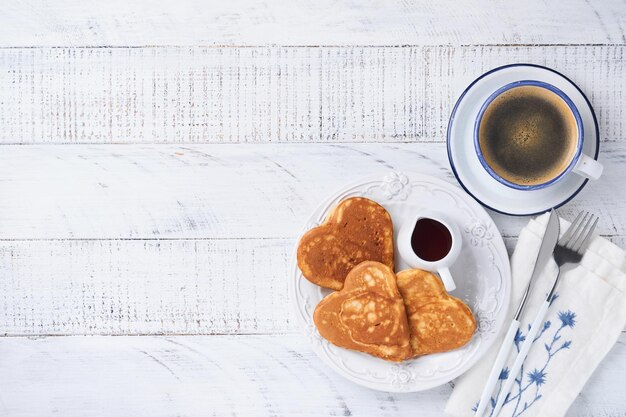 Pfannkuchen mit Beerenmarmelade und Honig in Form von Herzen und heißer Tasse Kaffee auf weißem Holzhintergrund. Konzeptfrühstück zum Valentinstag oder angenehme Überraschung für einen geliebten Menschen Tisch von oben betrachtet