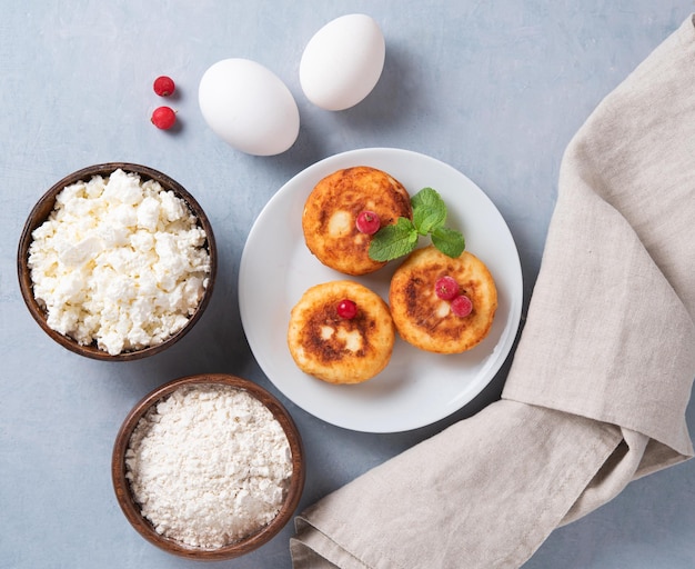 Pfannkuchen mit Hüttenkäse mit frischen Beeren und Sauerrahm auf blauem Hintergrund mit Eiern und Mehl das Konzept eines gesunden und leckeren Morgenfrühstücks