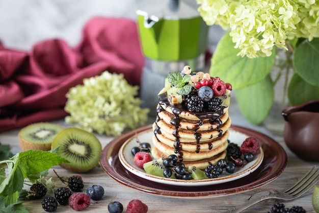 Pfannkuchen mit Schokoladensauce mit Beeren und Früchten mit einem Bouquet von Hortensien auf dem Tisch. Licht