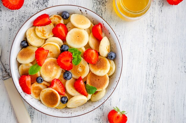 Pfannkuchen Müsli, Mini Pfannkuchen in einer Schüssel mit frischen Erdbeeren, Blaubeeren und Honig. Trend Essen. Horizontal, Draufsicht, Kopierraum.