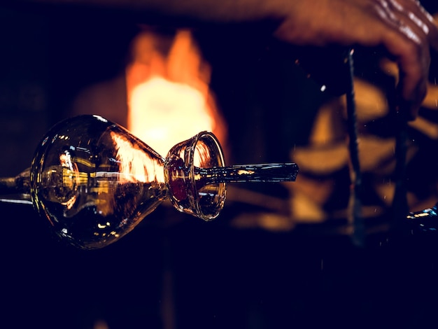 Pfeife mit glänzender handgefertigter Glasflasche in der Nähe einer brennenden Feuerflamme in einer dunklen Glasbläserwerkstatt