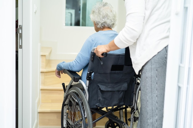 Pflegekräfte helfen asiatischen oder älteren alten Frauen, die im Rollstuhl sitzen, die Treppe im Haus hinauf zu stützen.