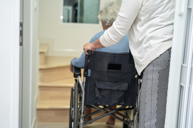 Pflegekräfte helfen asiatischen oder älteren alten Frauen, die im Rollstuhl sitzen, die Treppe im Haus hinauf zu stützen.