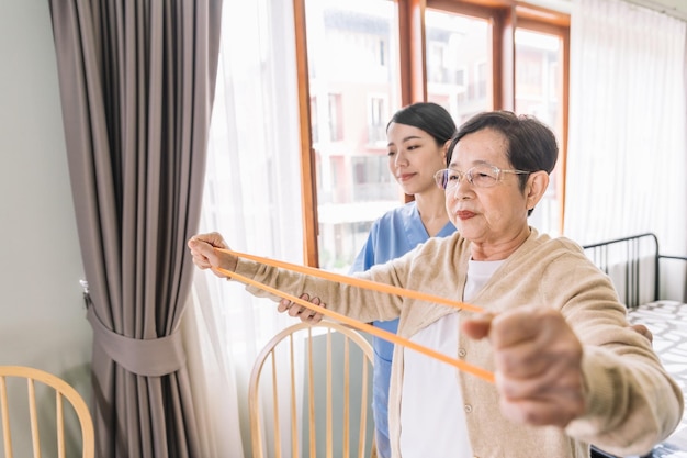 Pflegekraft, die Scrubs trägt, trainiert mit einer älteren asiatischen Frau, indem sie Widerstandsbandübungen für den älteren Patienten in der Physiotherapie-Behandlung verwendet Häusliches Gesundheits- und Pflegeheimkonzept