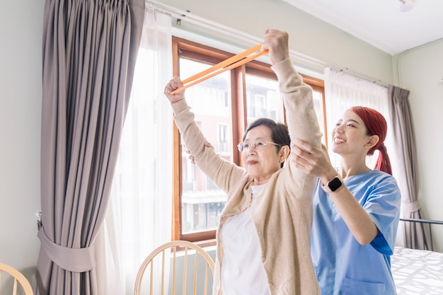 Pflegekraft, die Scrubs trägt, trainiert mit einer älteren asiatischen Frau, indem sie Widerstandsbandübungen für den älteren Patienten in der Physiotherapie-Behandlung verwendet Häusliches Gesundheits- und Pflegeheimkonzept