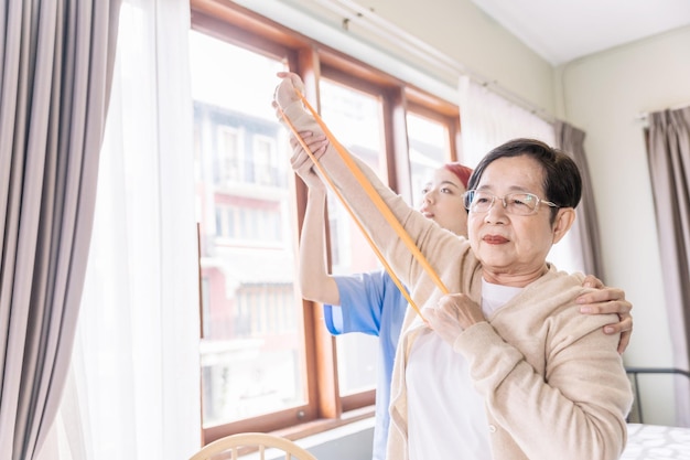 Pflegekraft, die Scrubs trägt, trainiert mit einer älteren asiatischen Frau, indem sie Widerstandsbandübungen für den älteren Patienten in der Physiotherapie-Behandlung verwendet Häusliches Gesundheits- und Pflegeheimkonzept